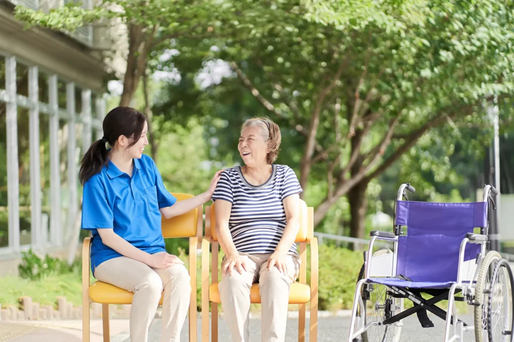 A smiling caregiver touches should of older woman as they sit outside in sunny weather