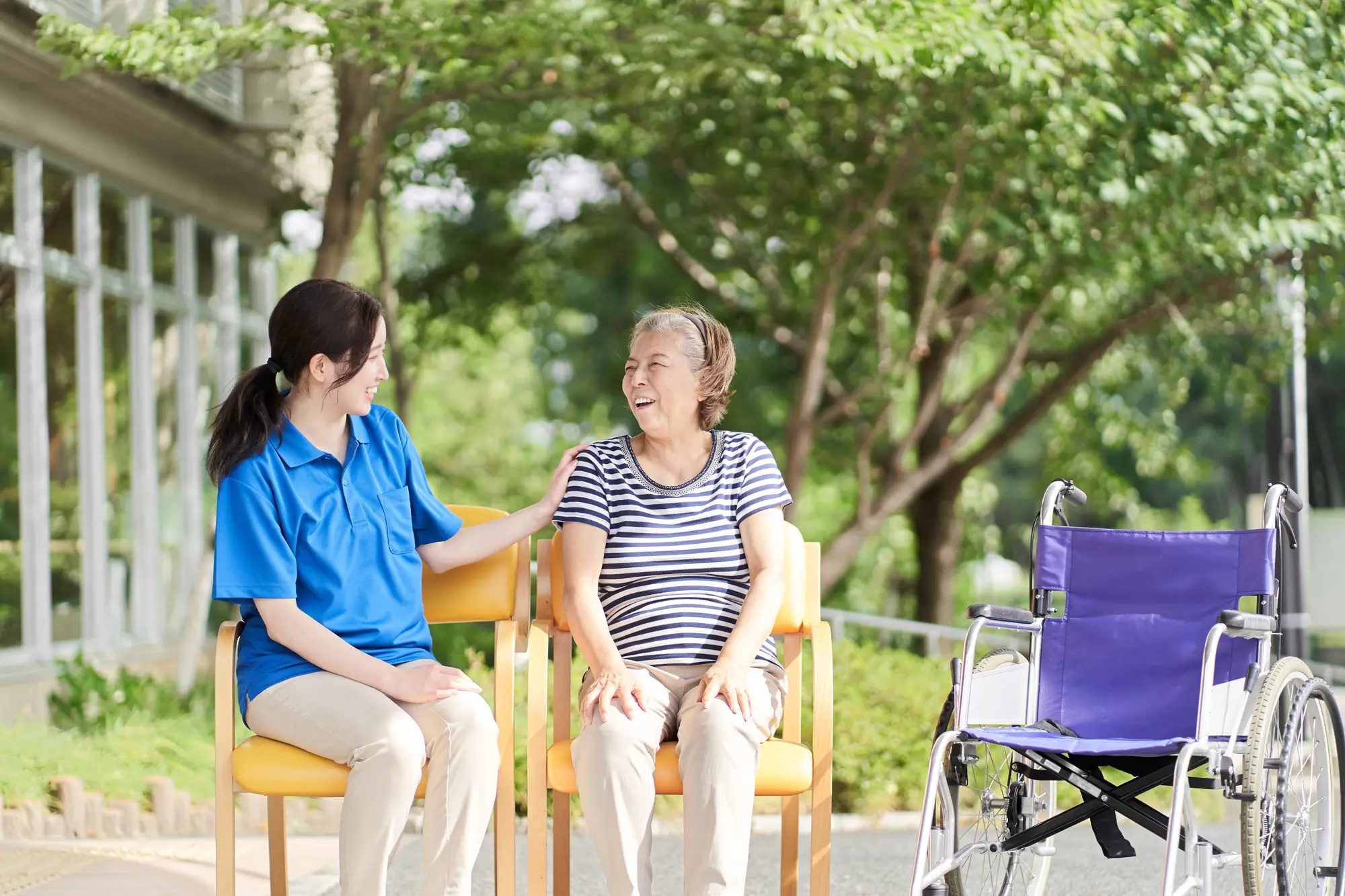 A smiling caregiver touches should of older woman as they sit outside in sunny weather