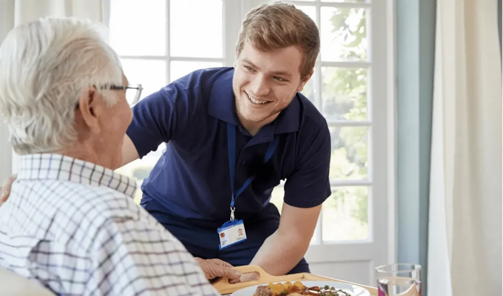 a caregiver smiles to an elderly man after serving him food