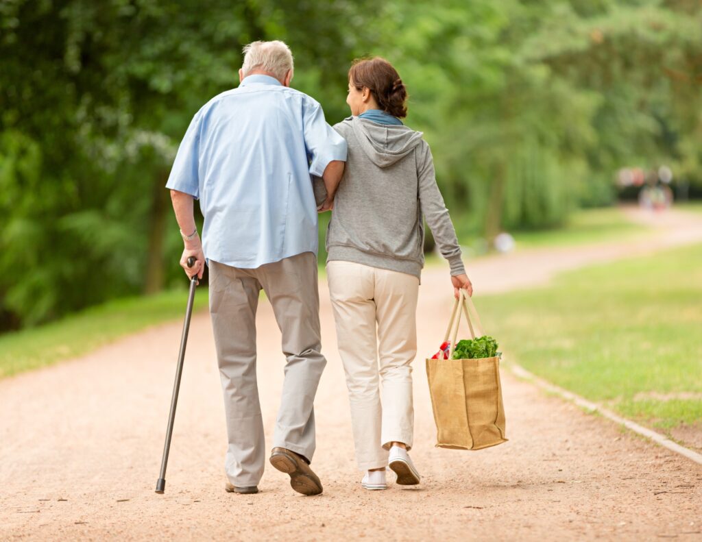 a caregiver helps a senior man walk home after they went grocery shopping
