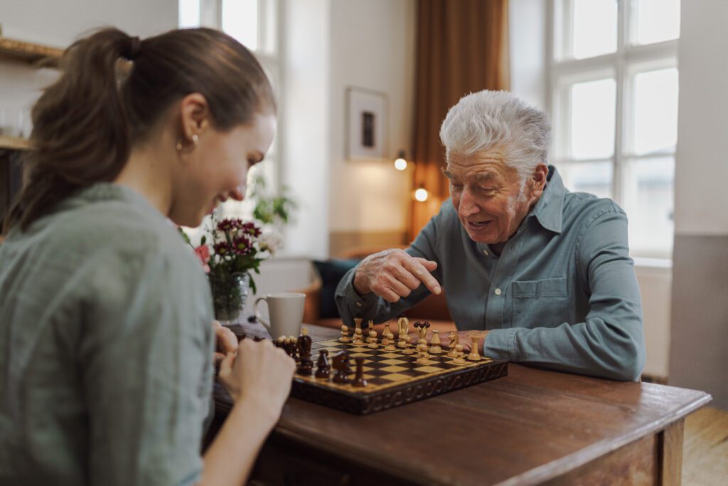 young woman plays chess with elderly man in home