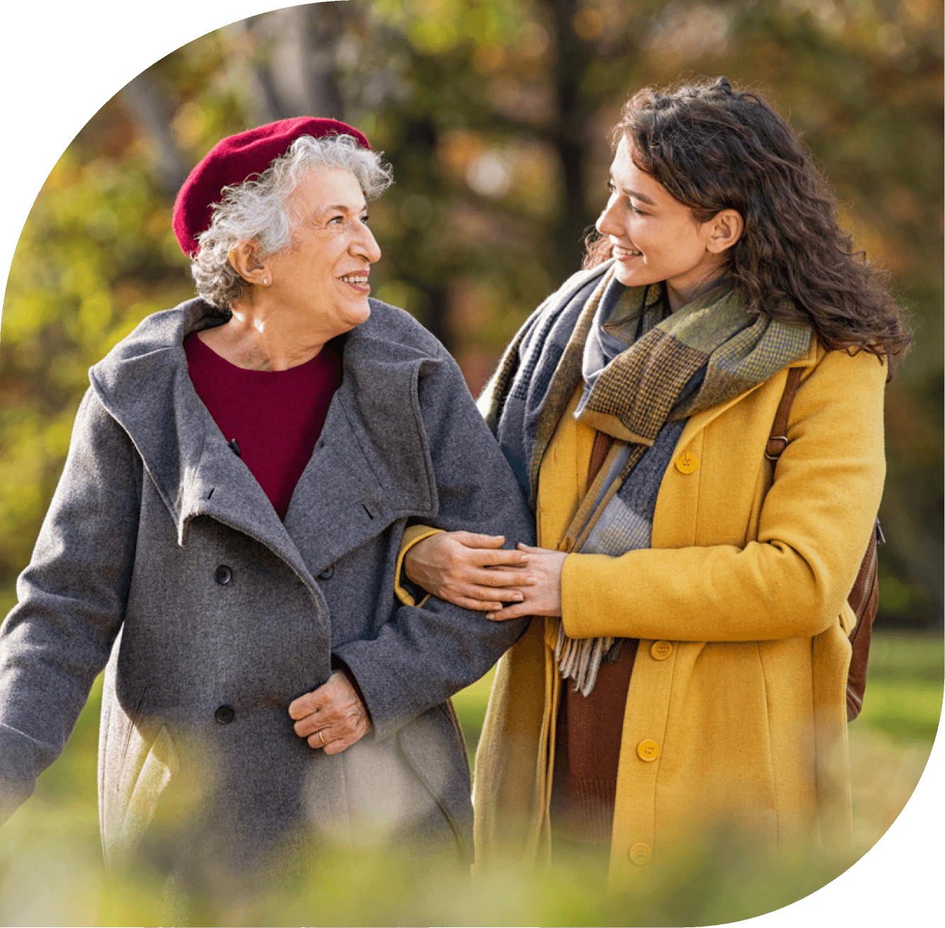 smiling young woman holds arm of elderly woman as they walk in the park