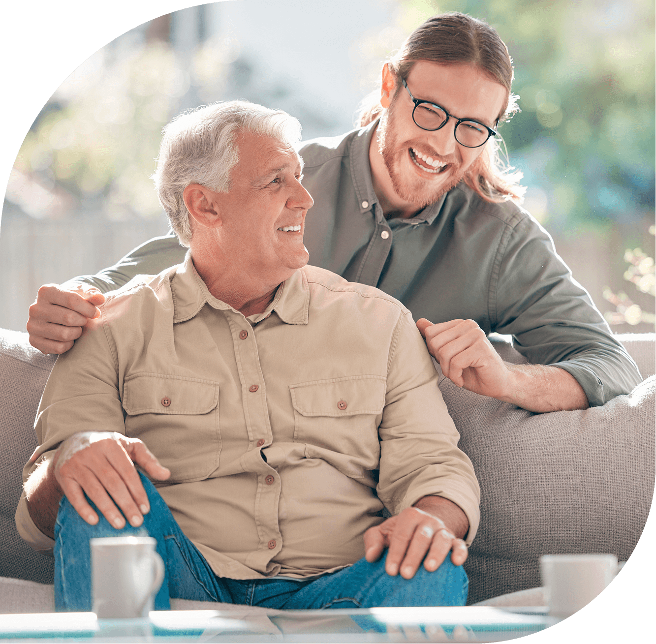 an smiling older man sits with a cup of coffee while a younger man stands behind him with hand on shoulder smiling