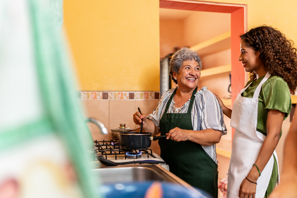 mature woman and younger woman in kitchen wearing aprons and smiling over pot cooking on stove