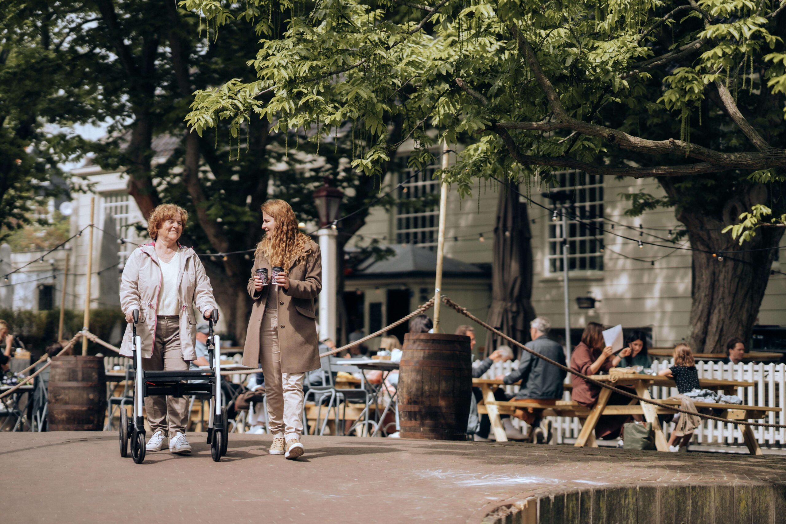 woman carries two to-go coffees while walking through town park with woman using walker