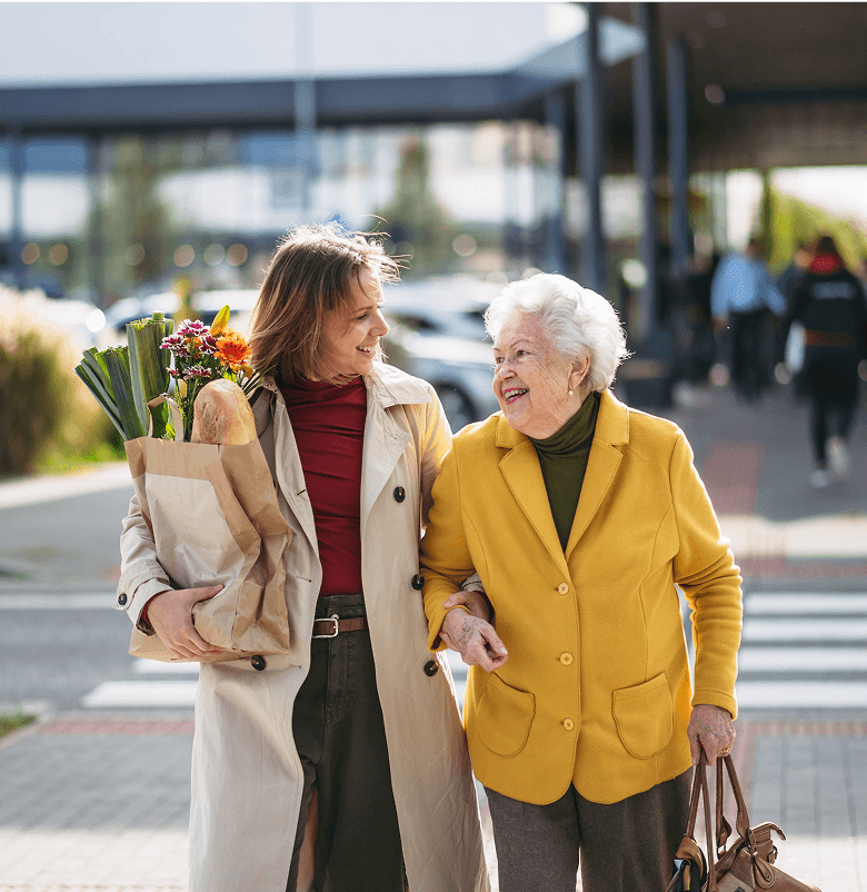 middle aged woman carries groceries for mature woman as they walk away from a shopping area