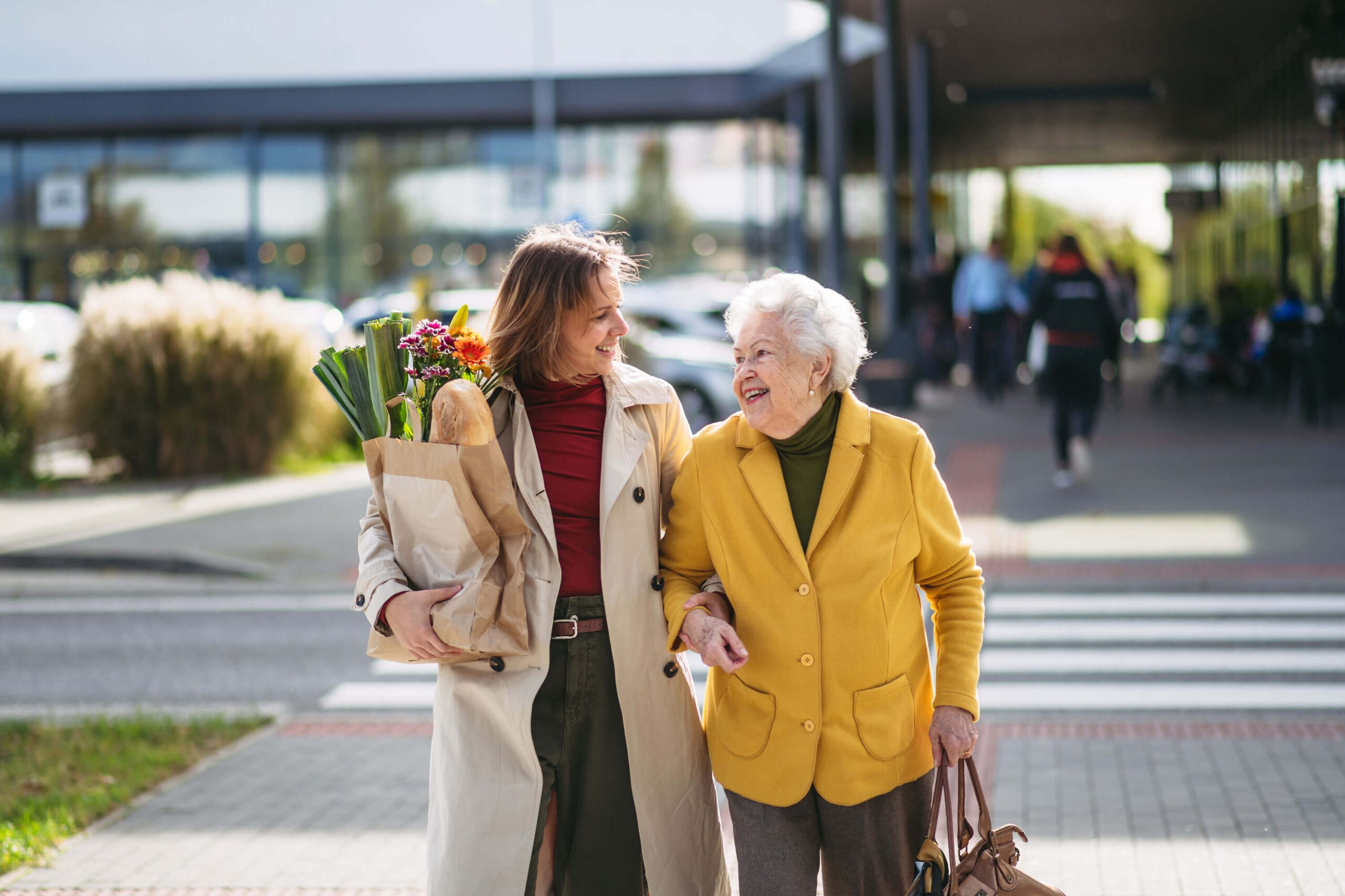 middle aged woman carries groceries for mature woman as they walk away from a shopping area