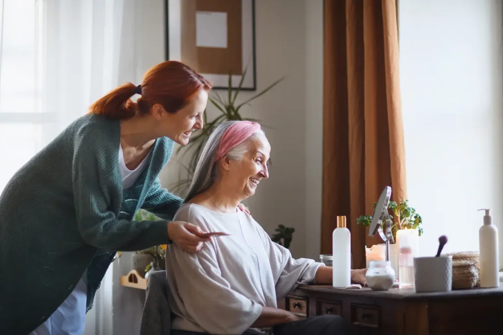 elderly woman smiles into mirror at younger woman holding hairbrush, who stands over her smiling back