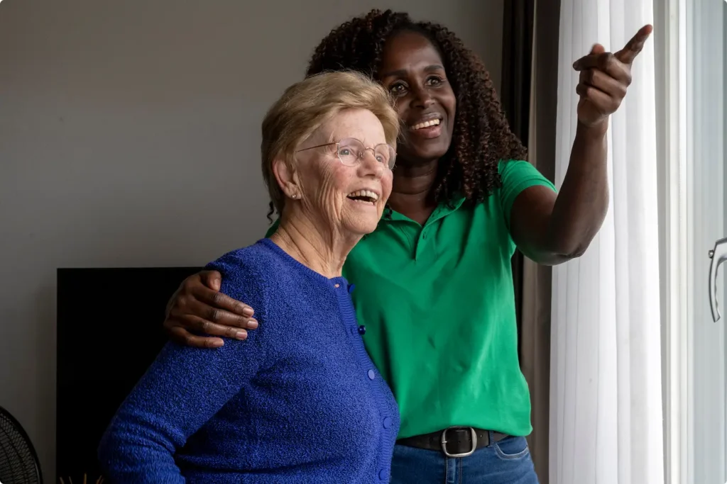 a middle aged woman puts her arm around an older woman while pointing out a window and smiling
