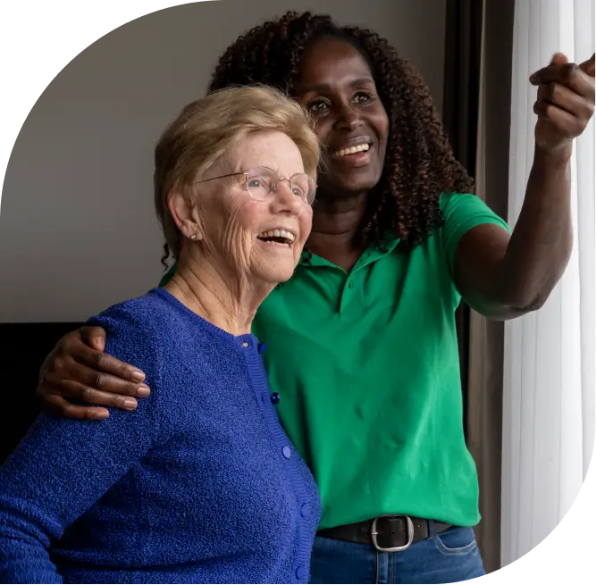 caregiver wraps arm around shoulder of senior woman as they point and look out a window smiling